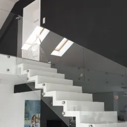 Nowoczesne białe schody wspornikowe z balustradą ze szkła hartowanego, mocowaną punktowo do stopni, na tle czarnej ściany i białego salonu z wiszącym fotelem.