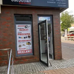 Fasada budynku Centrum Usługowego Szymanski z otwartymi drzwiami, szyldem reklamowym i plakatem informującym o dorabianiu kluczy, wyrobie pieczątek, grawerowaniu, pucharach i reklamie.