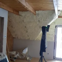 Poddasze w trakcie izolacji pianą PUR, widoczne drewniane belki stropowe, folia aluminiowa i narzędzia, odzież robocza na statywie.