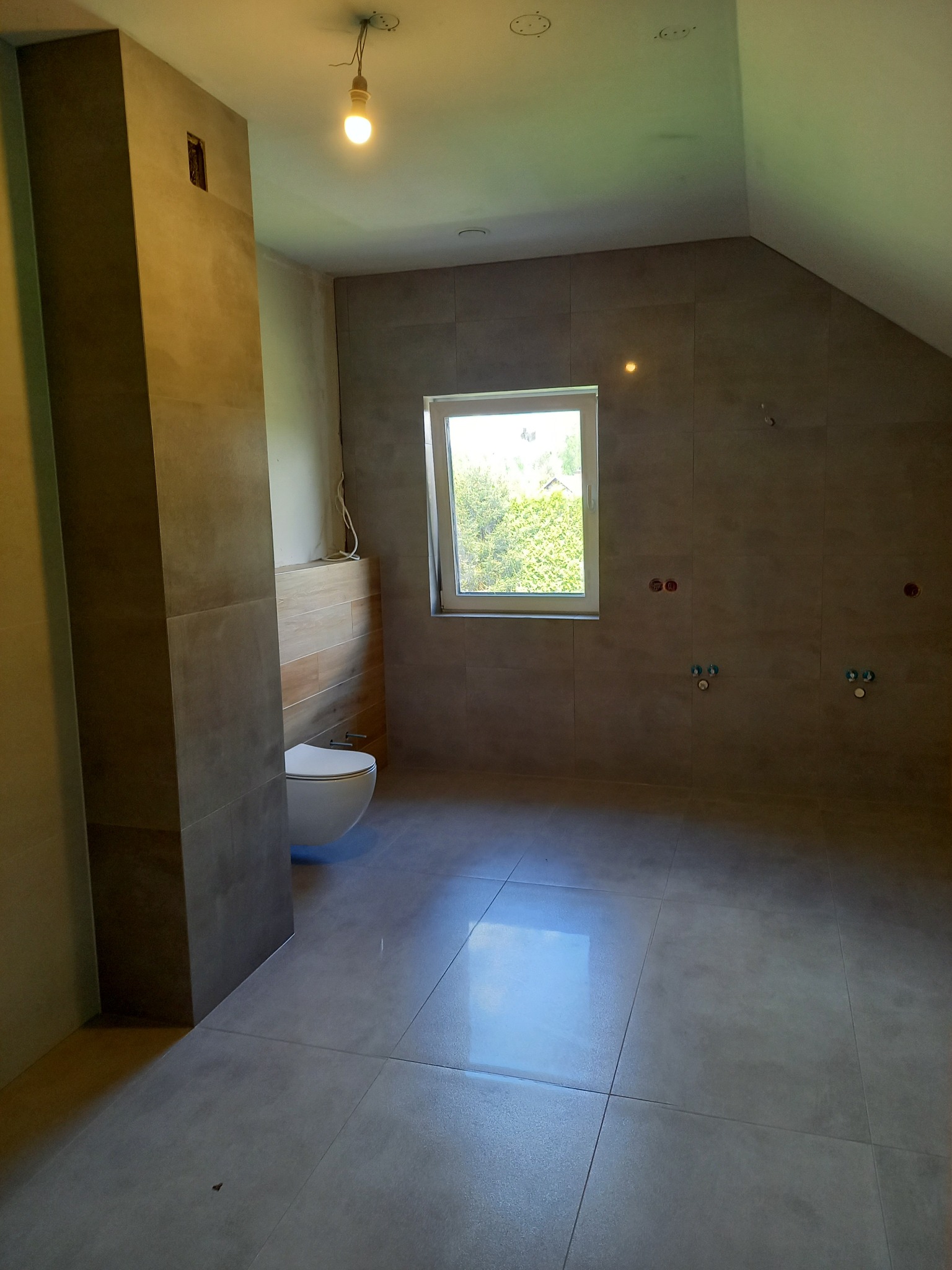 Surowy stan łazienki w Boguchwale po położeniu szarych płytek ściennych i podłogowych, widoczne wyprowadzenia instalacji wodnej i elektrycznej, częściowo zamontowana toaleta wisząca.