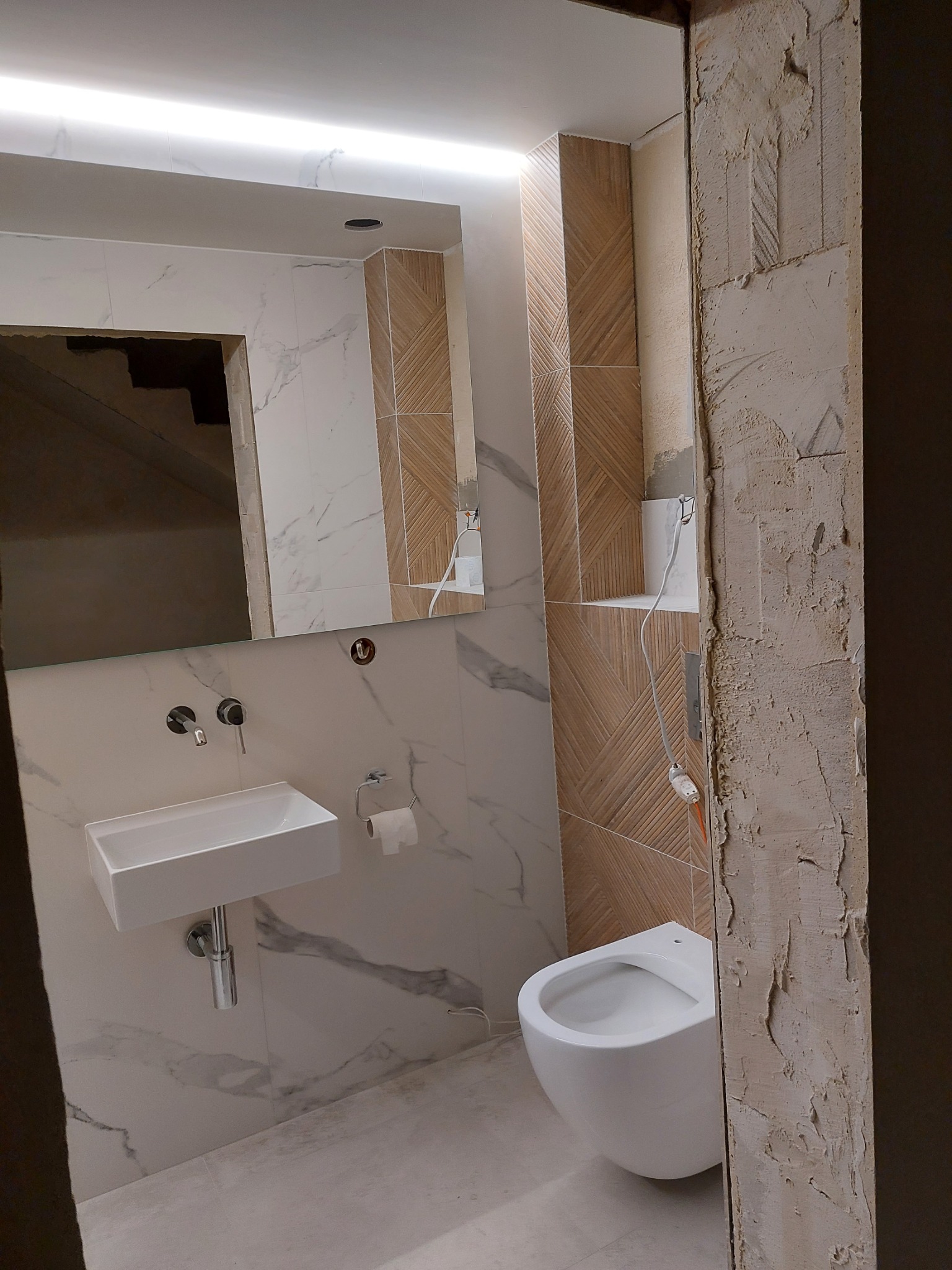 Fragment łazienki w trakcie prac: białe płytki marmurkowe, drewnopodobna mozaika na ścianie, podwieszana toaleta i umywalka, surowy ościeżnica drzwi.