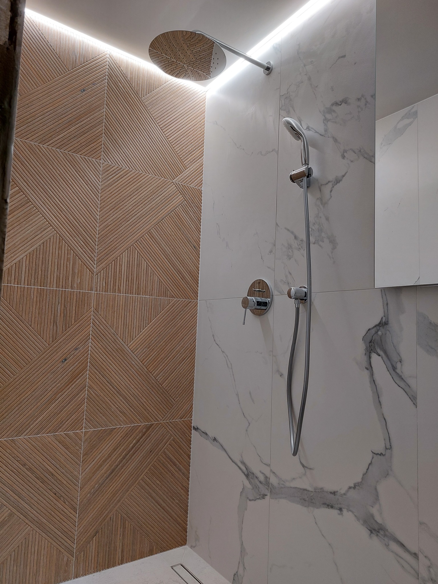 Nowoczesna kabina prysznicowa z mozaiką drewnopodobną i płytkami imitującymi marmur, oświetlenie LED pod sufitem, deszczownica i bateria termostatyczna z drewnianym akcentem.