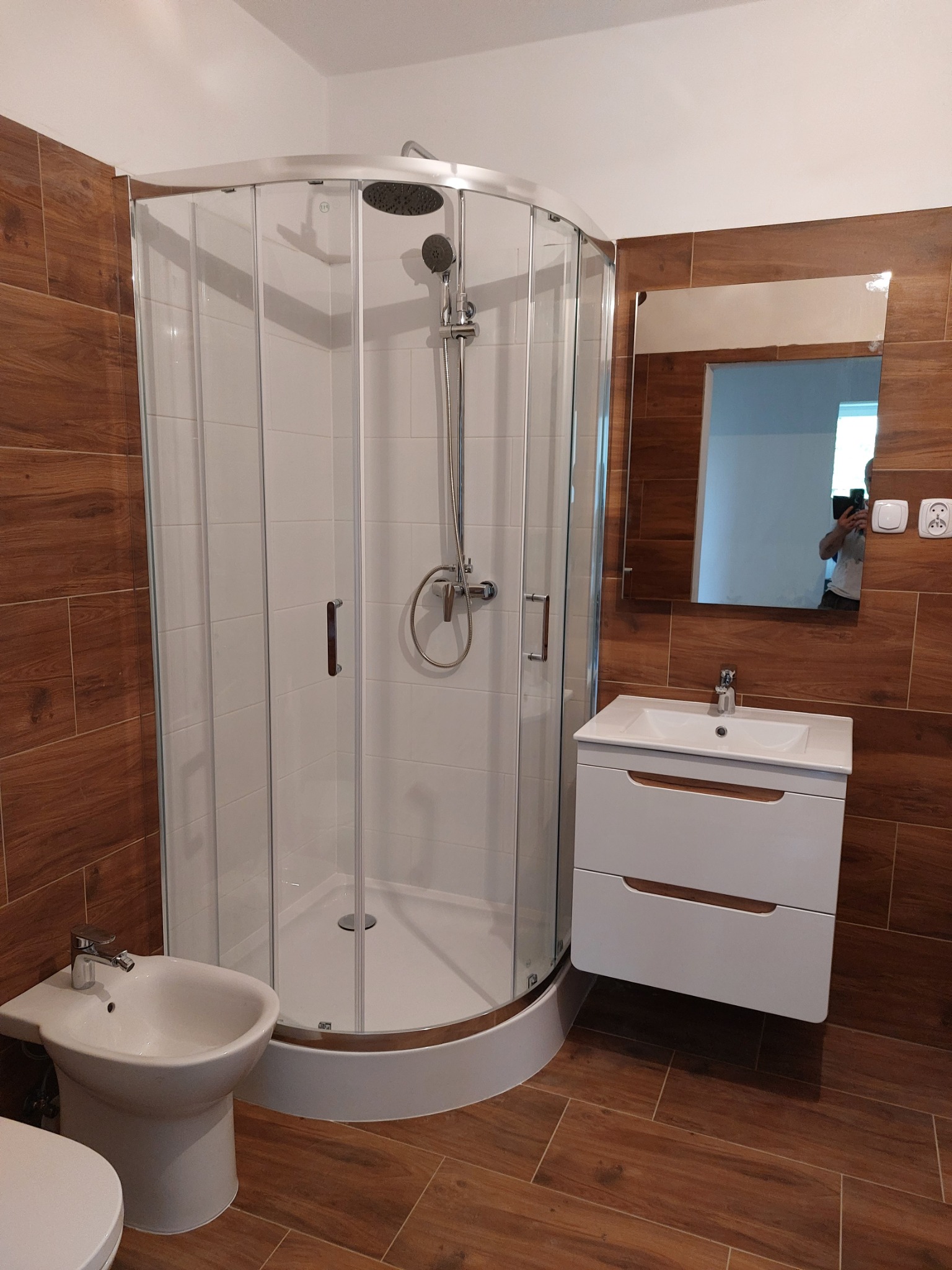 Nowoczesna łazienka z kabiną prysznicową narożną, białą szafką pod umywalkę z dwoma szufladami, bidetem i brązowymi płytkami imitującymi drewno na ścianach i podłodze. W lustrze odbija się osoba...