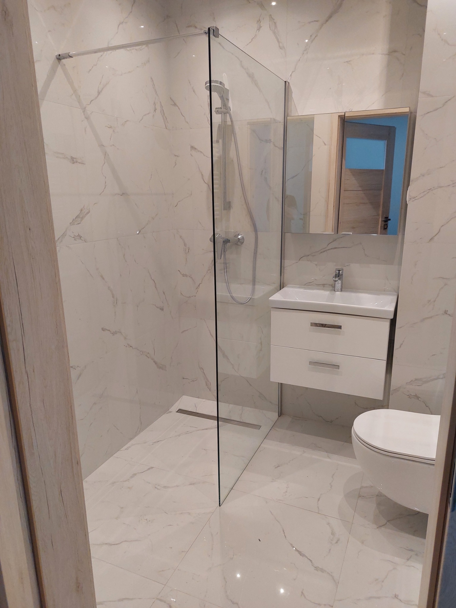 Nowoczesna łazienka z kabiną prysznicową typu walk-in, marmurowymi płytkami ściennymi i podłogowymi, wiszącą szafką z umywalką i podwieszaną toaletą.