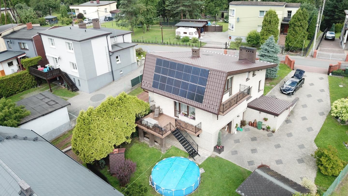 Widok z góry na dom jednorodzinny z brązowym dachem, na którym zamontowane są panele fotowoltaiczne; obok domu zaparkowany czarny samochód z otwartym bagażnikiem, a w ogrodzie rozłożony basen.
