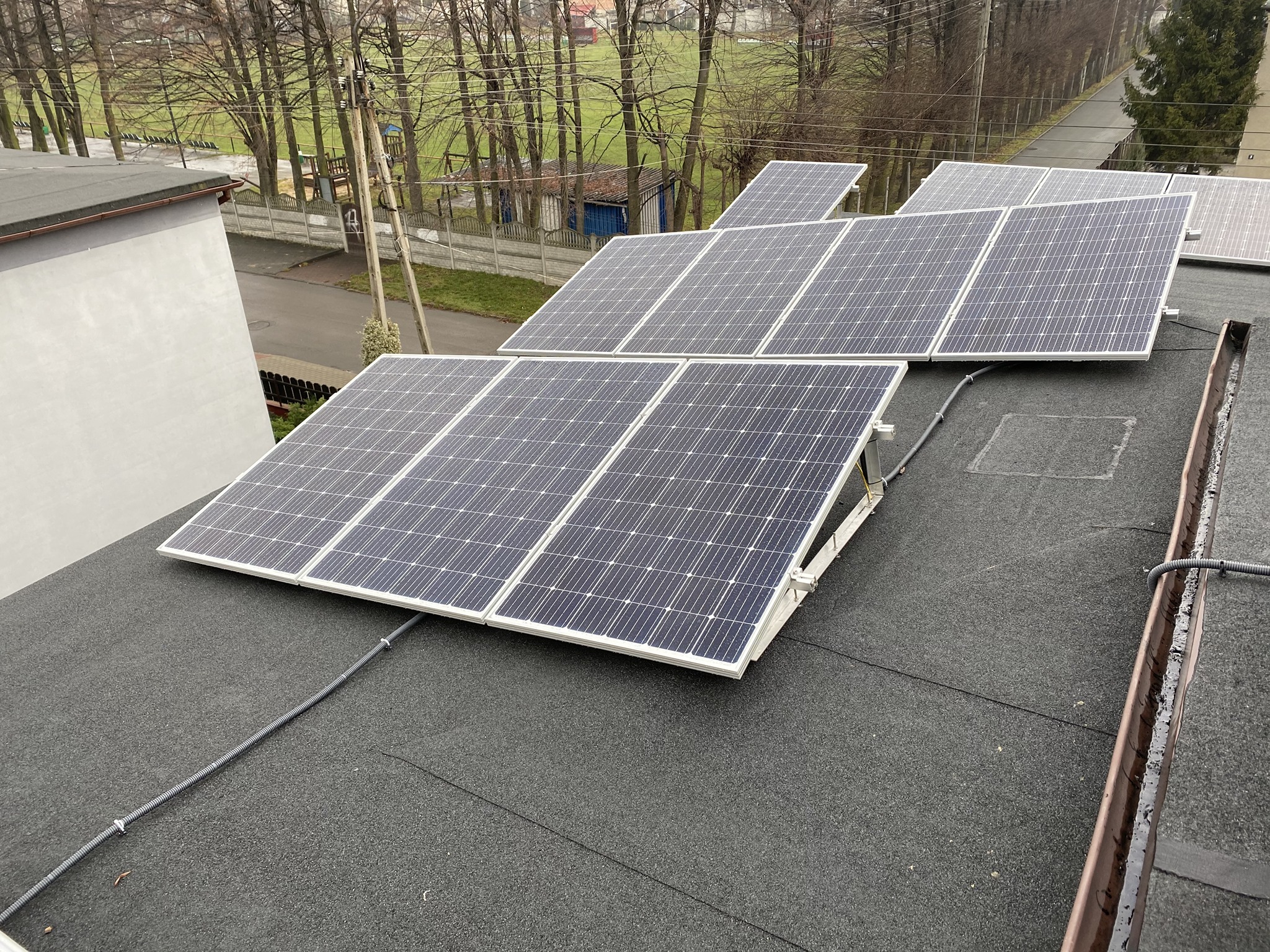 Instalacja paneli słonecznych na płaskim, bitumicznym dachu budynku z widocznymi przewodami i otoczeniem miejskim w tle.