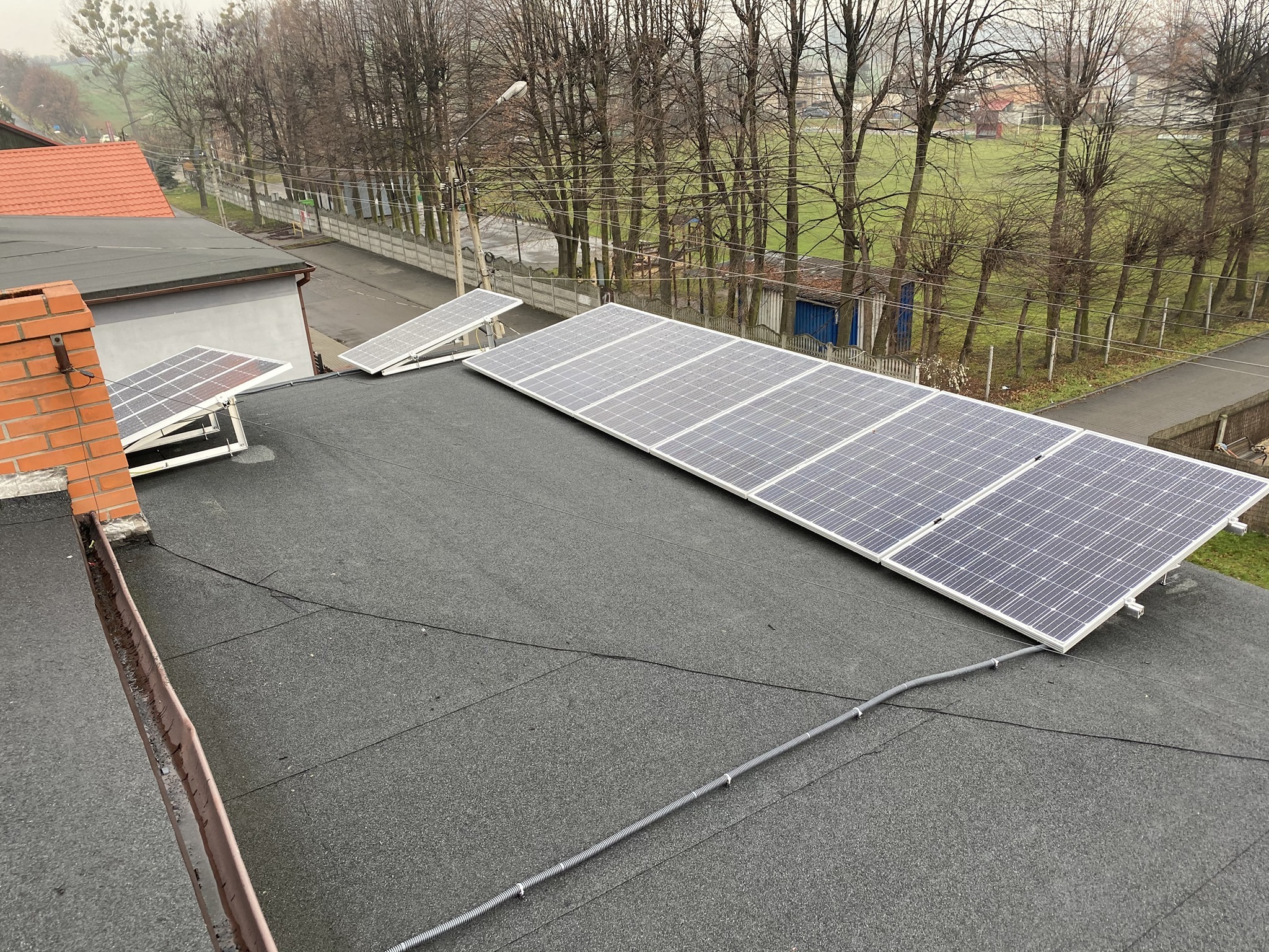 Instalacja paneli słonecznych na płaskim dachu budynku z widocznym kominem z cegły i otoczeniem w tle. Widoczne kable prowadzące od paneli.