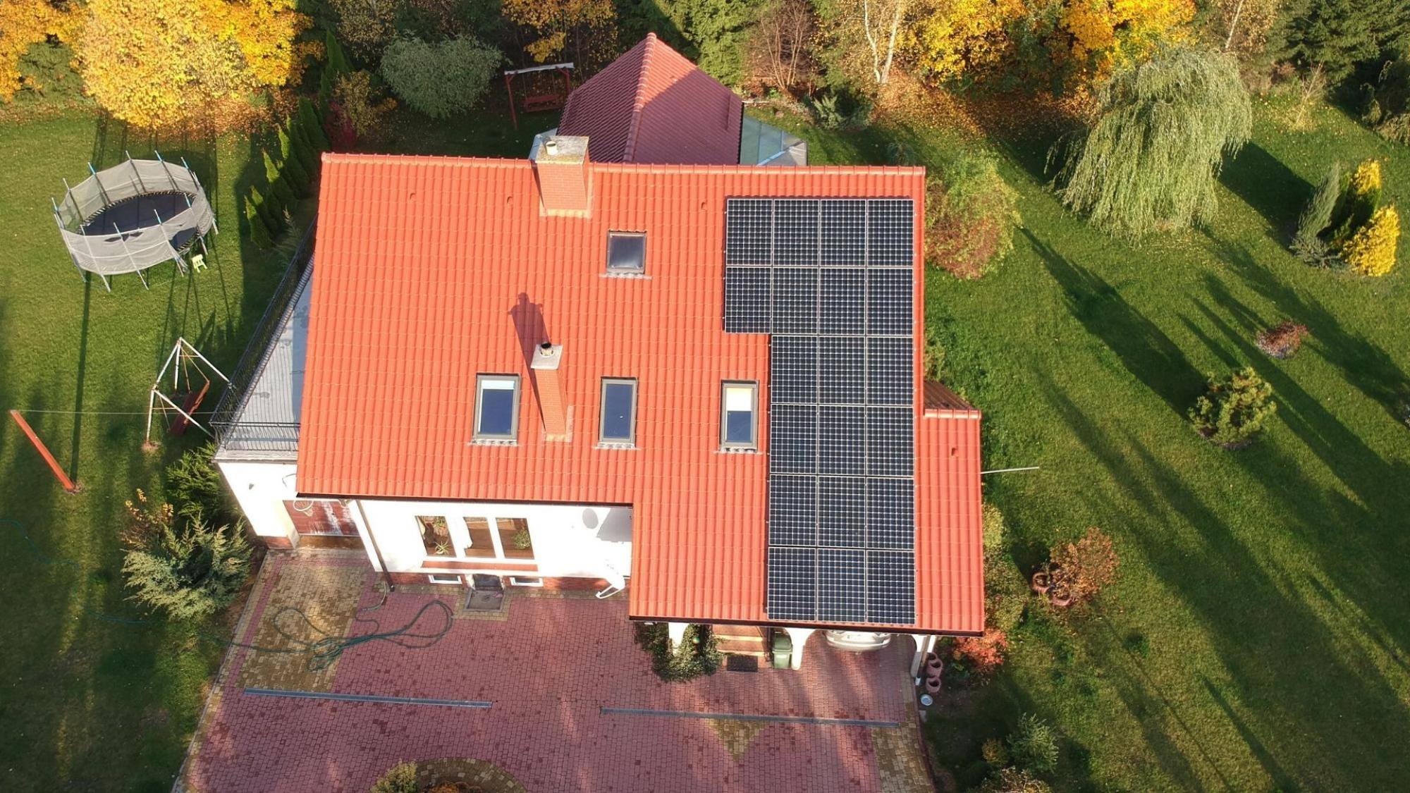Widok z góry na dom z czerwoną dachówką, na której zamontowano panele słoneczne w prostokątnym układzie, częściowo zacienione przez komin. W tle widoczny ogród z trampoliną i drzewami w jesiennych...