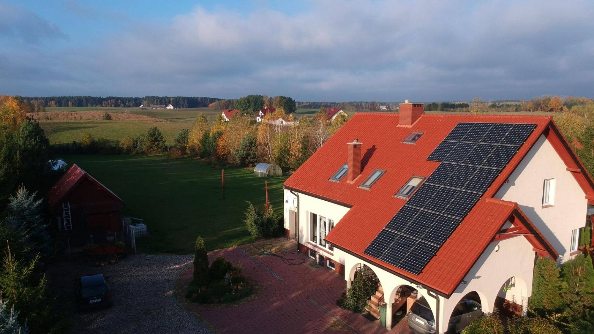 Widok z góry na dom jednorodzinny z czerwoną dachówką, na której zamontowane są panele słoneczne. W tle rozległe pola i lasy, błękitne niebo z delikatnymi chmurami. Na podjeździe przed domem...
