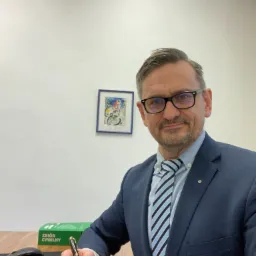 Portret mężczyzny w garniturze i okularach, siedzącego przy biurku i podpisującego dokument złotym piórem, z zielonym pudełkiem z napisem 'Zbiór Cywilny' w tle.