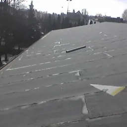 Szary dach z papy z widocznymi łączeniami pasów, na którym leży poziomica i trójkątny kawałek papieru z taśmą klejącą, w tle drzewa i budynki.