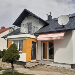 Nowoczesny dom jednorodzinny z białą elewacją, elementami w kolorze drewna, balkonem z ozdobną balustradą i markizą w biało-czerwone paski, widoczne schody wejściowe z palet.