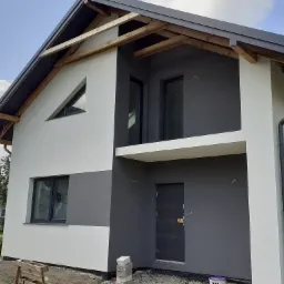 Nowoczesny dom jednorodzinny z dwukolorową elewacją w odcieniach szarości i bieli, widoczne drewniane elementy konstrukcji dachu, okna z folią ochronną, przed wejściem ułożone bloczki betonowe...