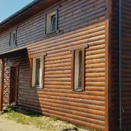 Dom z bali pomalowany na brązowo, widoczne okna z białymi ramami i metalowa konstrukcja ganku.