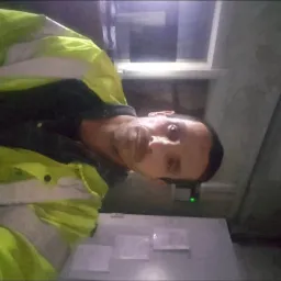 Mężczyzna w odblaskowej kurtce pozuje do selfie w pomieszczeniu z białą szafką i widocznymi dokumentami.