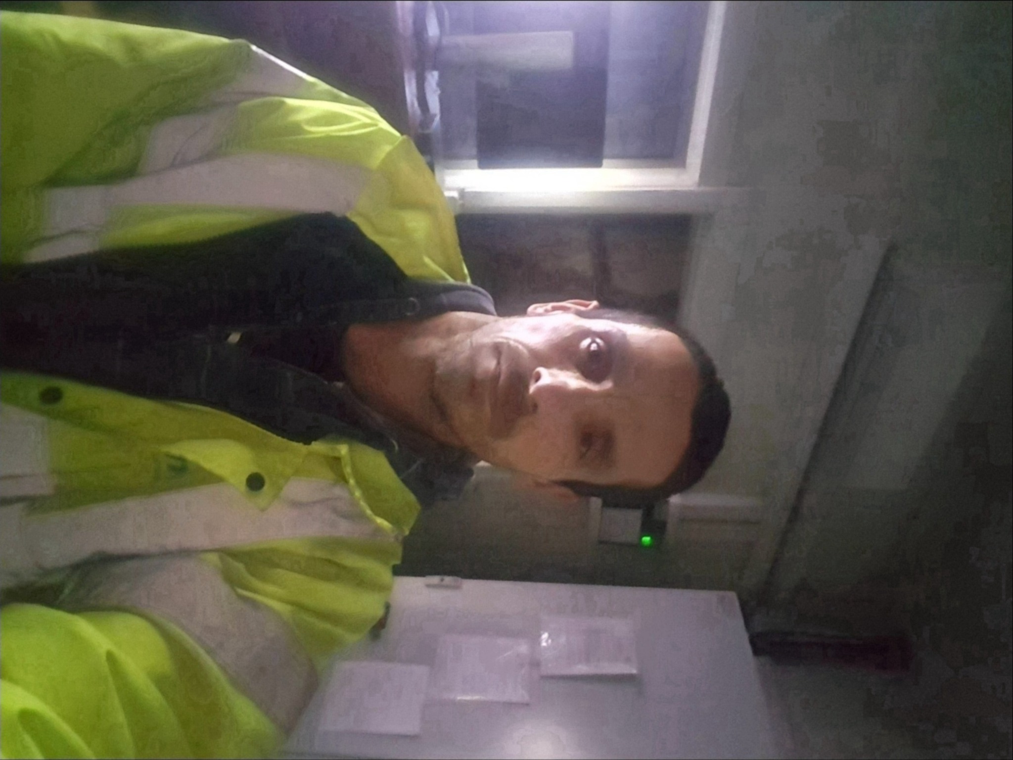 Mężczyzna w odblaskowej kurtce pozuje do selfie w pomieszczeniu z białą szafką i widocznymi dokumentami.