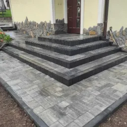 Schody wejściowe do domu wyłożone szarą kostką brukową, obrzeża z ciemniejszej kostki, w tle elewacja z kamienną dekoracją.