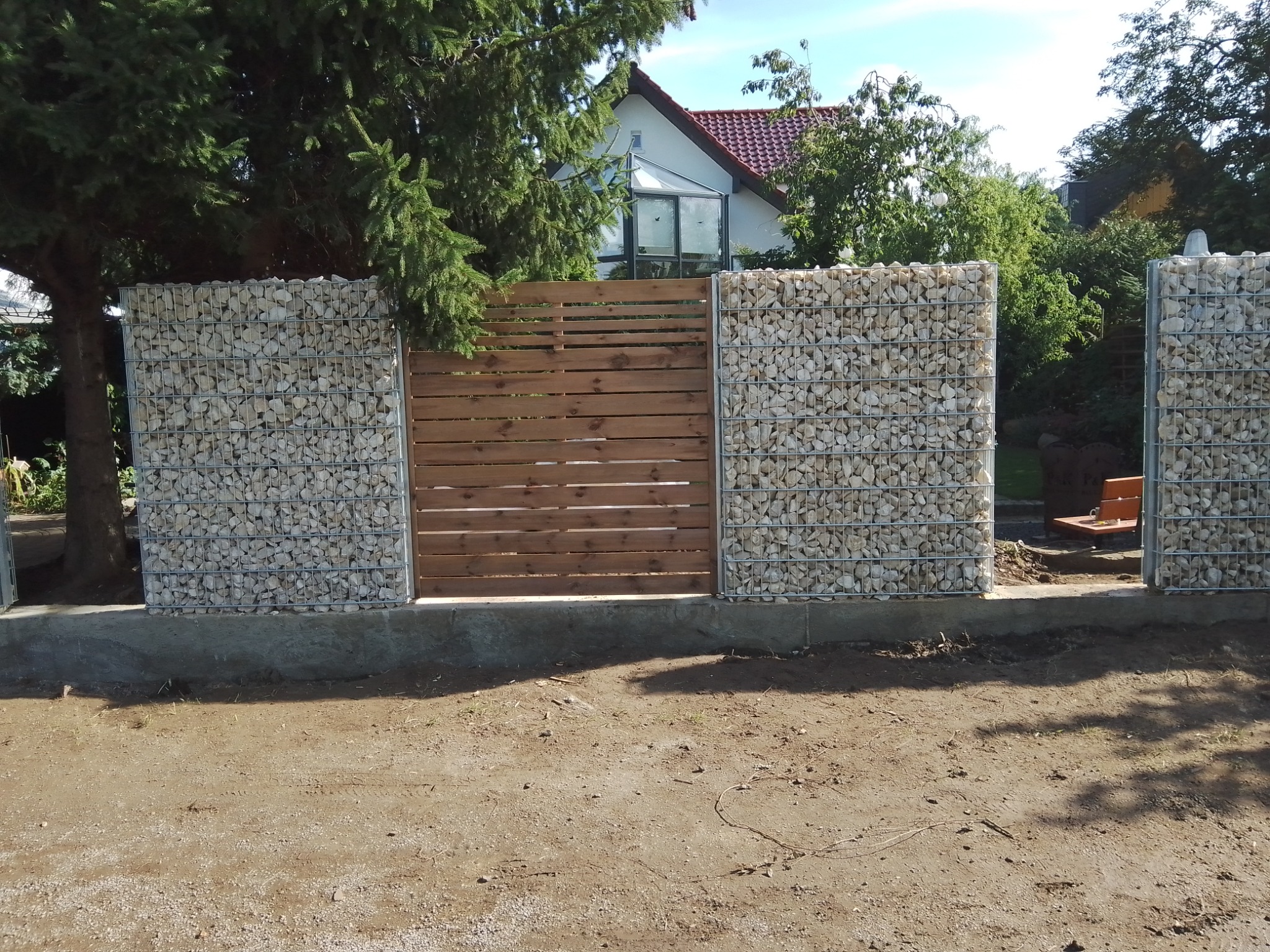 Nowoczesne ogrodzenie z gabionów wypełnionych białym kamieniem i drewnianych paneli, widoczne na tle zieleni i domu z czerwoną dachówką.