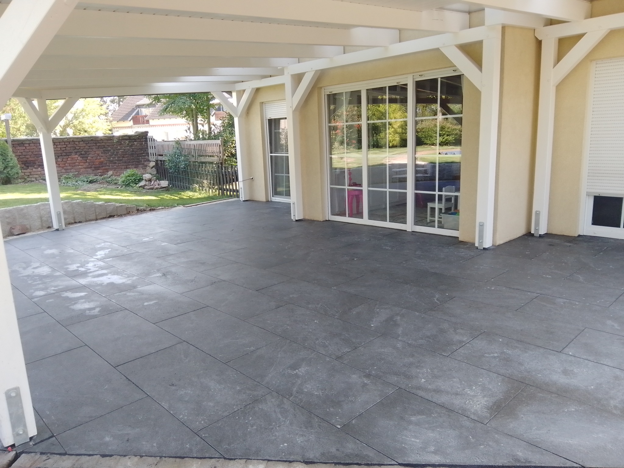 Zadaszony taras z szarymi, prostokątnymi płytkami ułożonymi w regularny wzór, widok na ogród przez duże, przeszklone drzwi przesuwne.