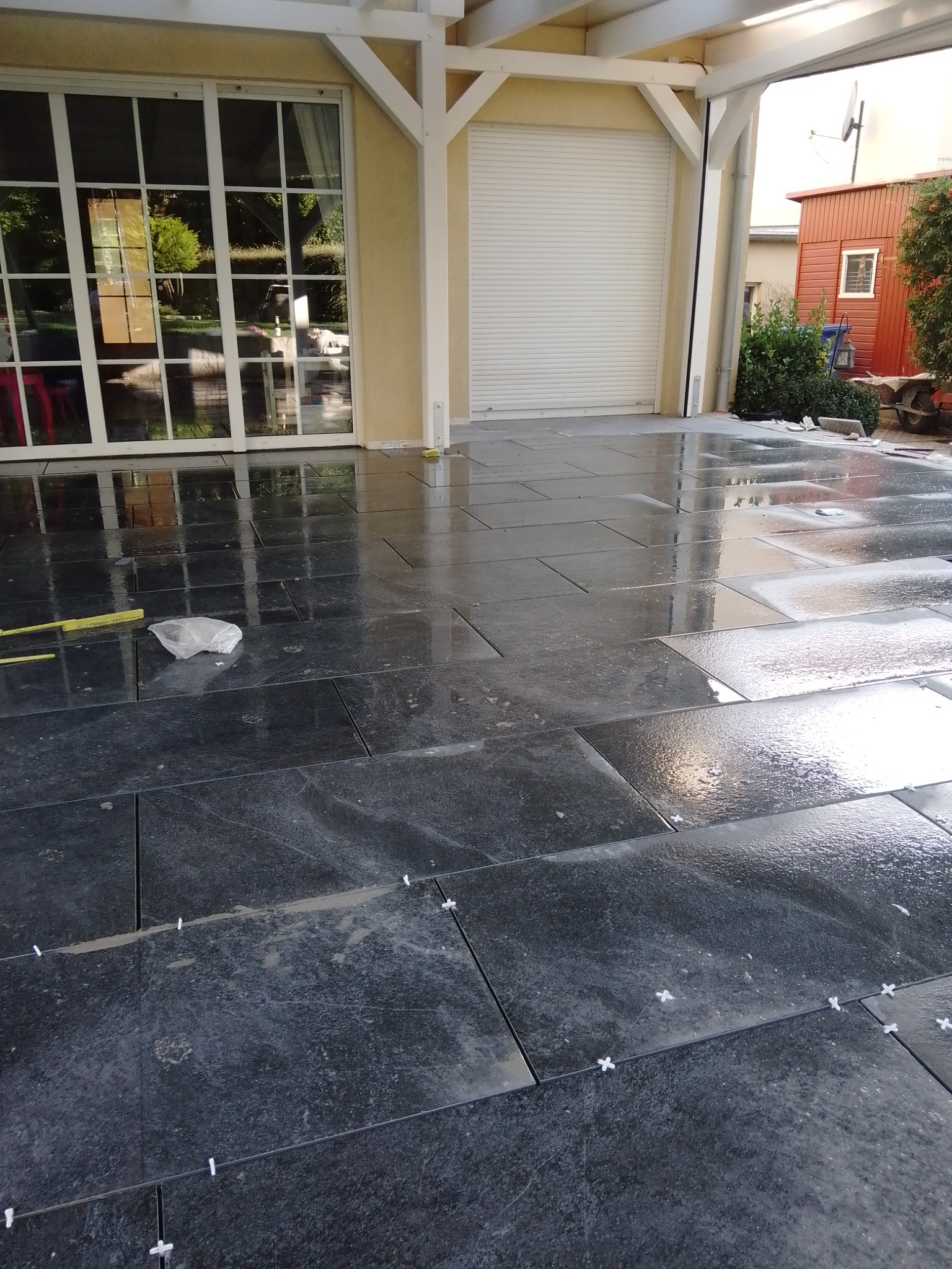 Układanie ciemnych płytek na tarasie z widocznymi krzyżykami dystansowymi, mokra powierzchnia odbija światło, w tle budynek z dużymi oknami i roletą.