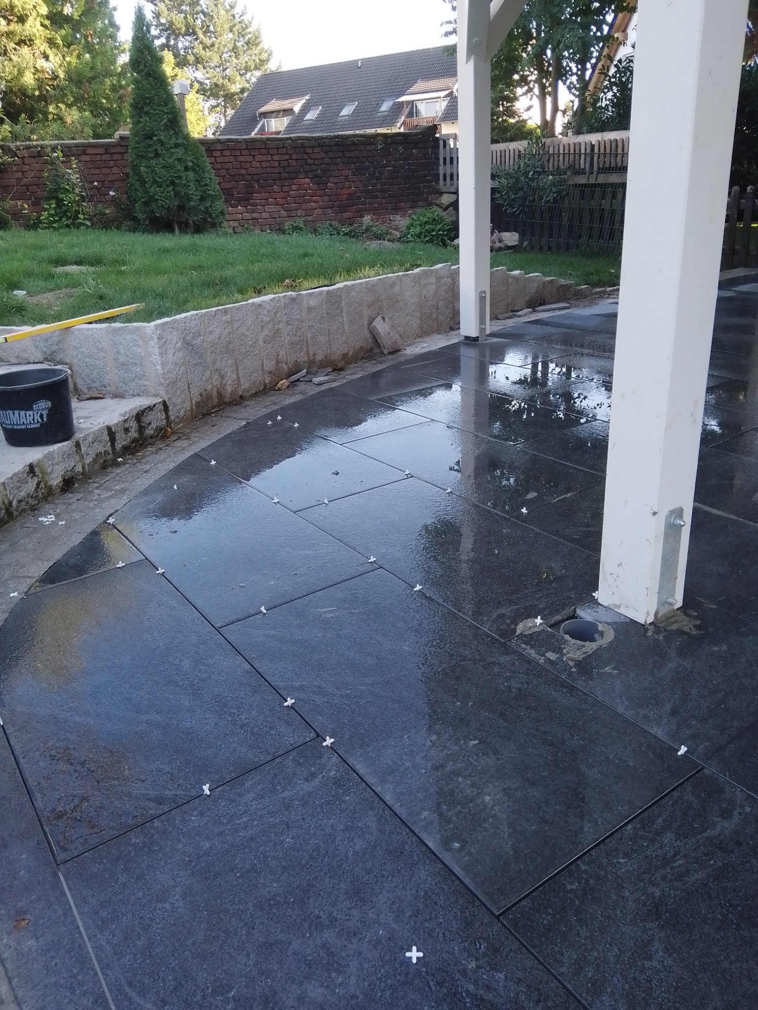 Układanie ciemnych, prostokątnych płytek na zewnątrz, z użyciem krzyżyków dystansowych, widoczny fragment ogrodu z murem oporowym i słupami konstrukcji.