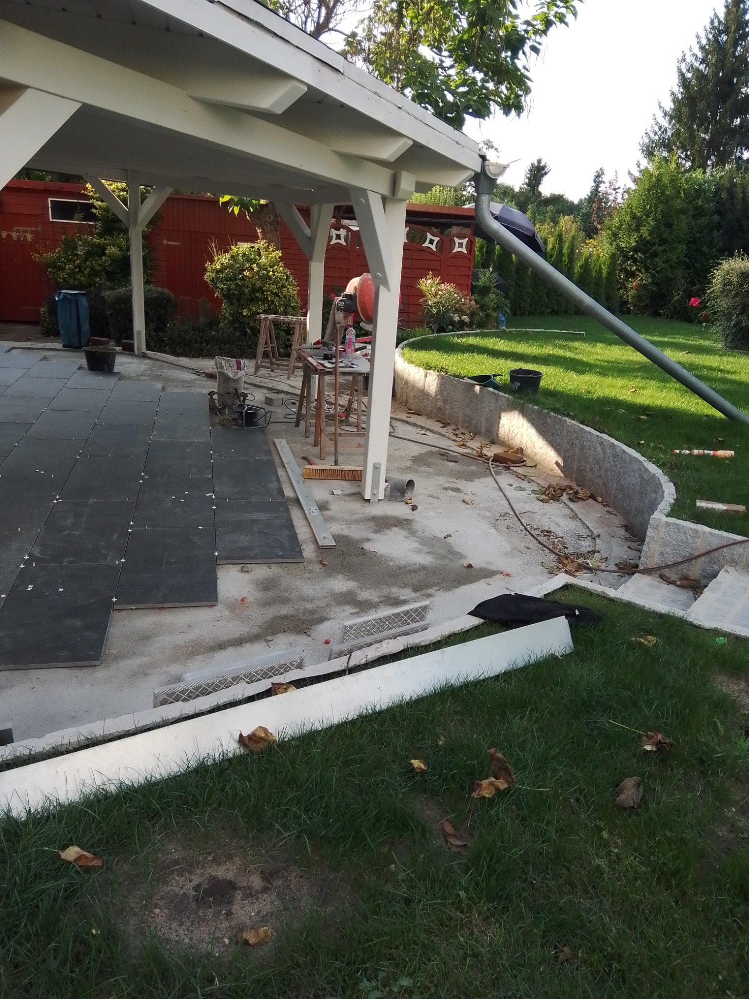 Układanie ciemnych płytek na tarasie pod zadaszeniem, widoczny mikser do zaprawy, poziomica i narzędzia, ogród w tle.