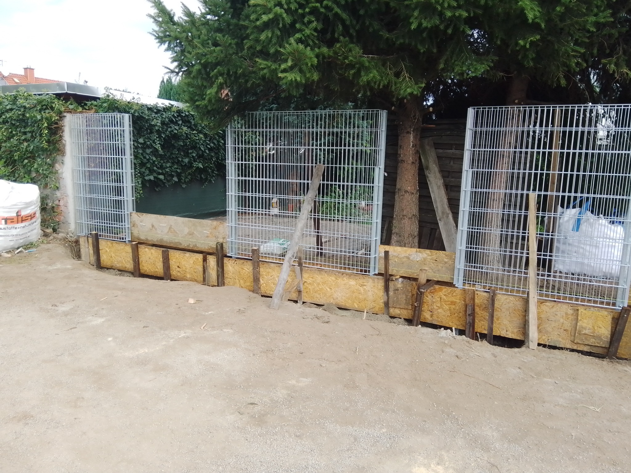 Przygotowanie szalunku i metalowych paneli do wylania fundamentu ogrodzenia na posesji, widoczne drewniane deski, metalowe panele ogrodzeniowe, worki z materiałem budowlanym w tle.