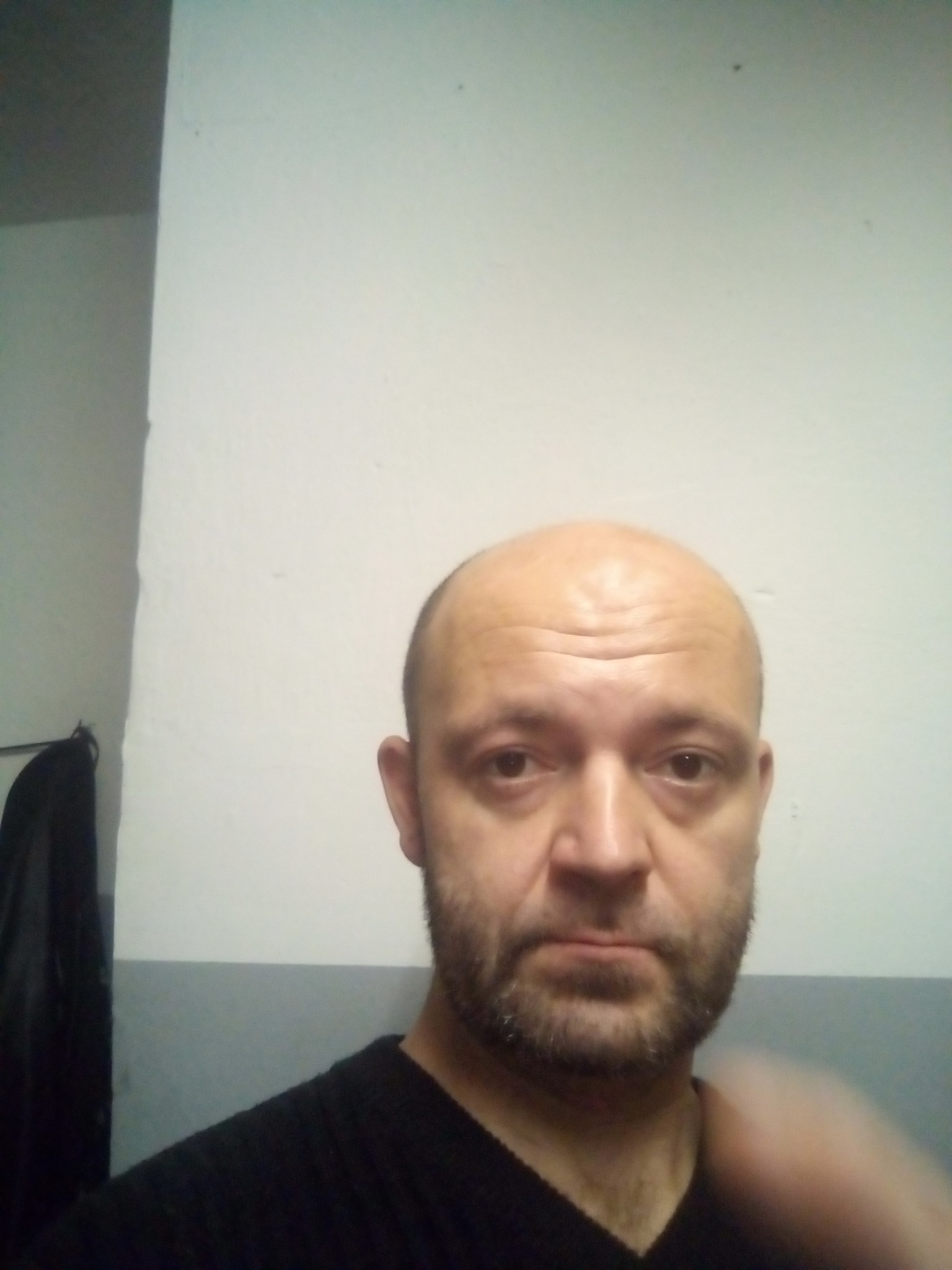 Mężczyzna z krótką brodą i łysą głową pozuje na tle ściany z szarym paskiem, ubrany w czarną koszulkę.