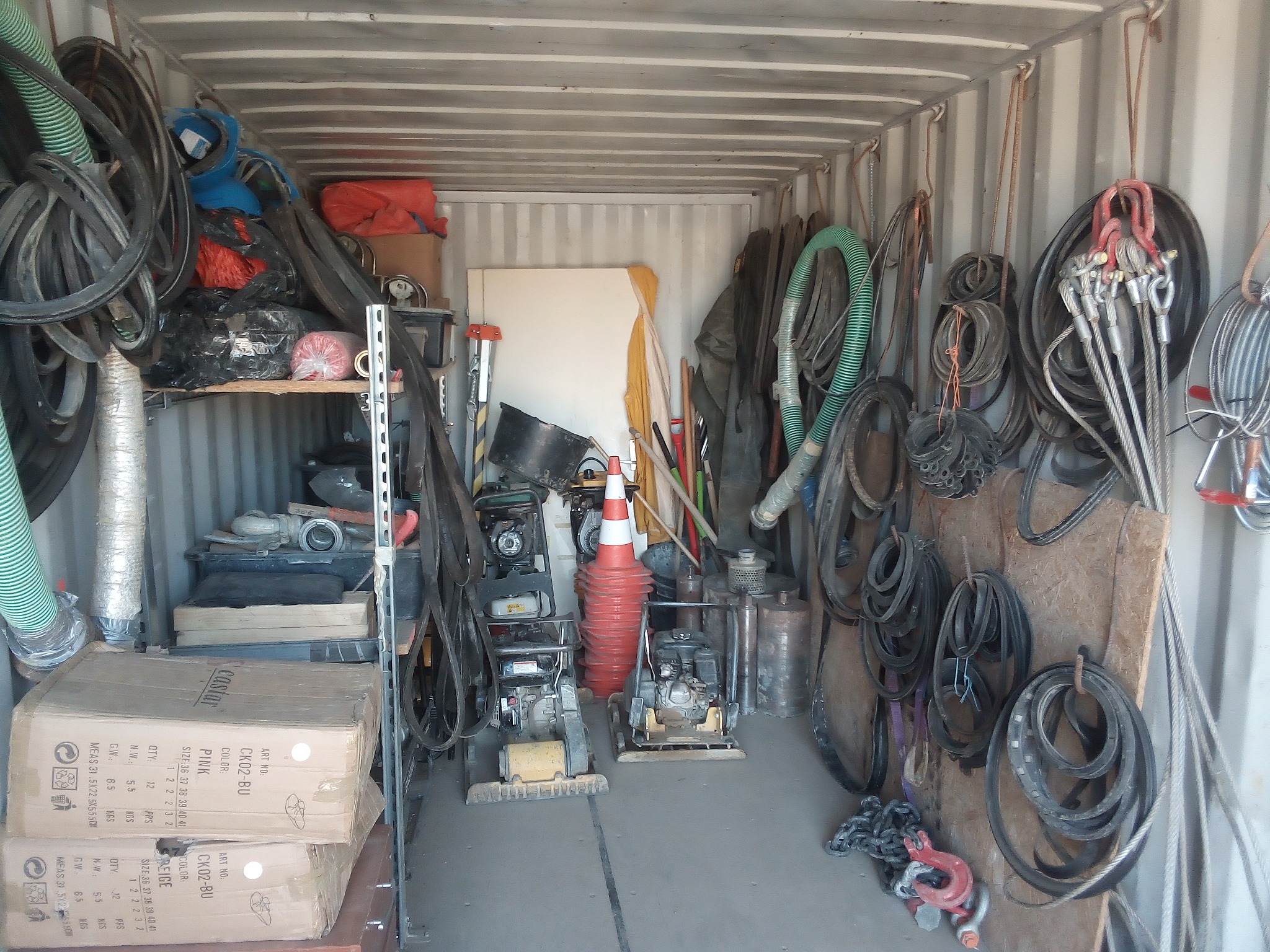 Wnętrze kontenera magazynowego wypełnione narzędziami budowlanymi, wężami przemysłowymi, pasami transportowymi, linami stalowymi i zagęszczarkami gruntu. Na półce kartony z obuwiem.