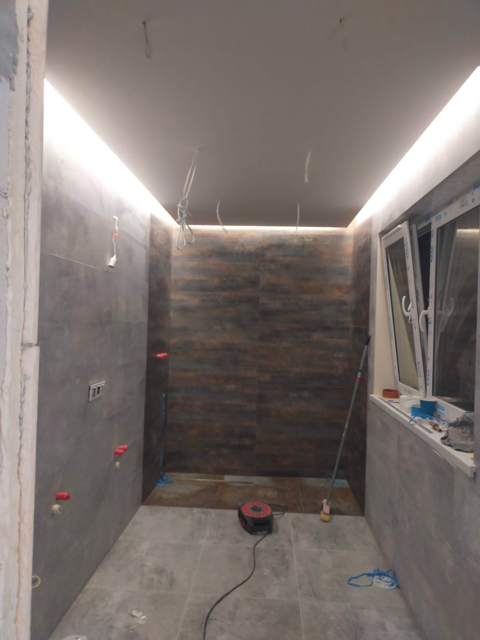 Wnętrze łazienki w trakcie remontu z szarymi płytkami na ścianach i podłodze, widoczne wystające kable, częściowo wykończona ściana z brązowymi akcentami, otwarte okno z akcesoriami na parapecie...