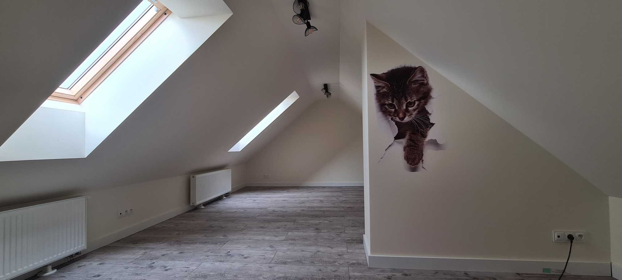 Poddasze po remoncie z panelami podłogowymi i naklejką kota na ścianie, widoczne okna dachowe i grzejnik.