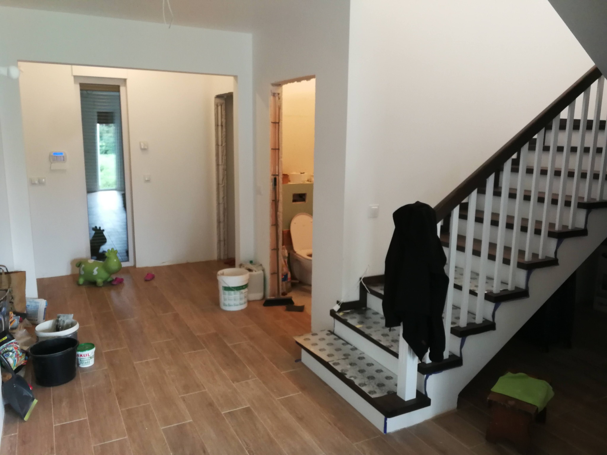 Wnętrze domu w trakcie prac wykończeniowych: brązowe panele podłogowe, białe ściany, schody z ciemnymi stopniami i białymi balustradami, widoczne wiadro z farbą i zabawki dziecięce, drzwi do łazienki.