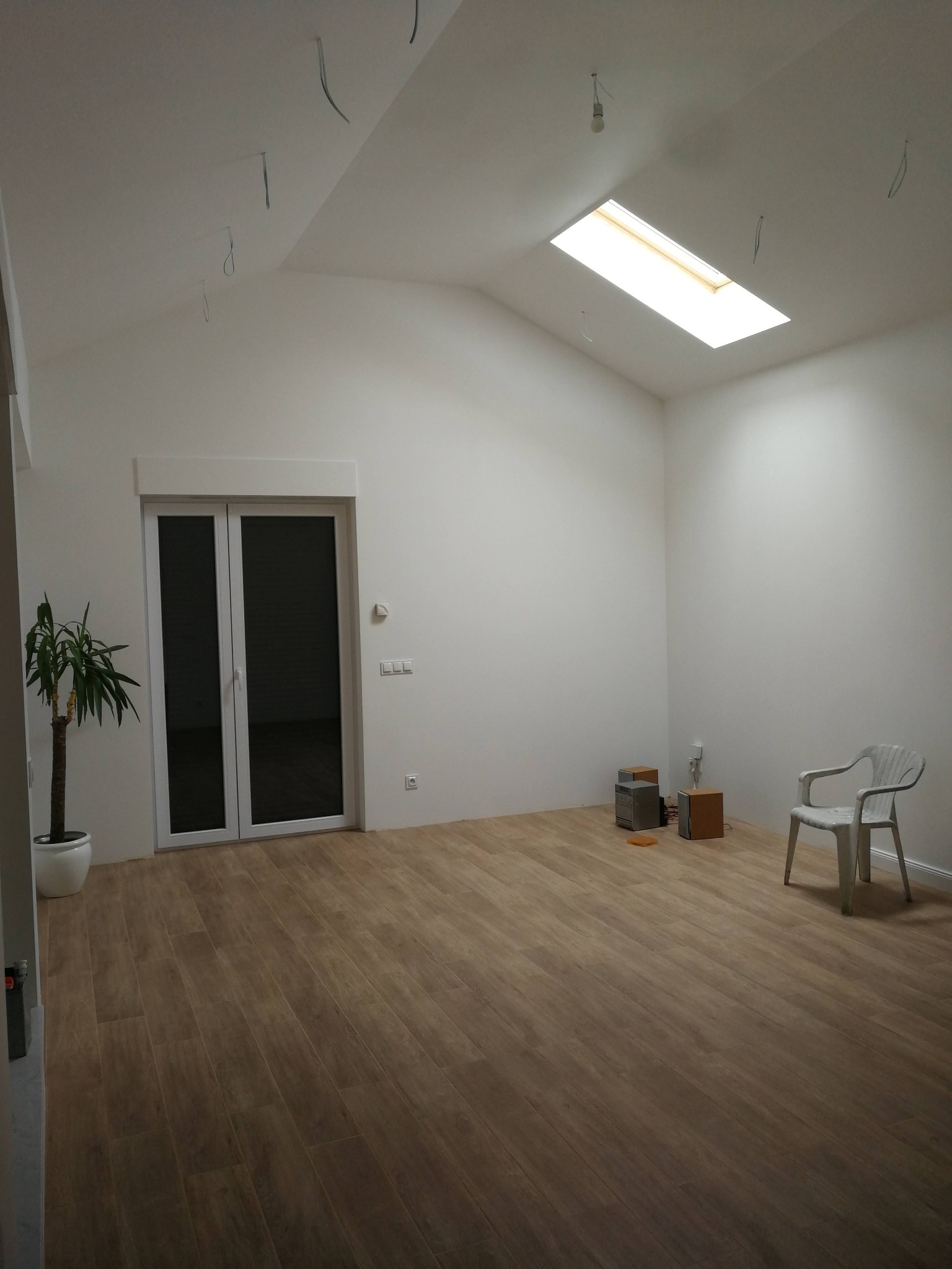 Wnętrze pokoju po remoncie w Poznaniu z nową podłogą panelową, gładkimi ścianami, oknem dachowym i przygotowanymi przewodami elektrycznymi do montażu oświetlenia. Widoczna roślina doniczkowa...