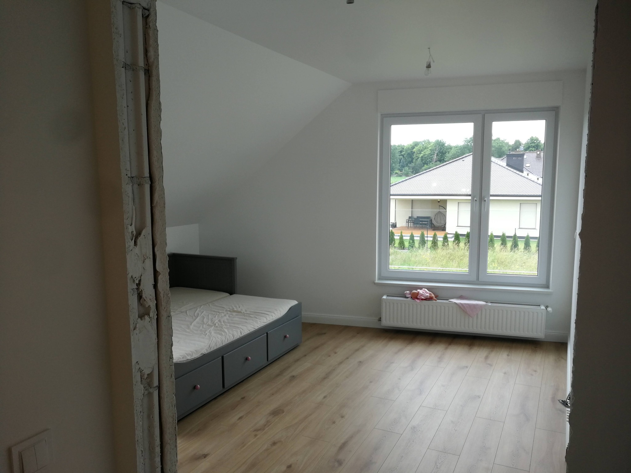 Pokój na poddaszu w trakcie prac wykończeniowych: widoczna surowa ościeżnica, łóżko z szufladami, grzejnik i panele podłogowe w jasnym odcieniu drewna.