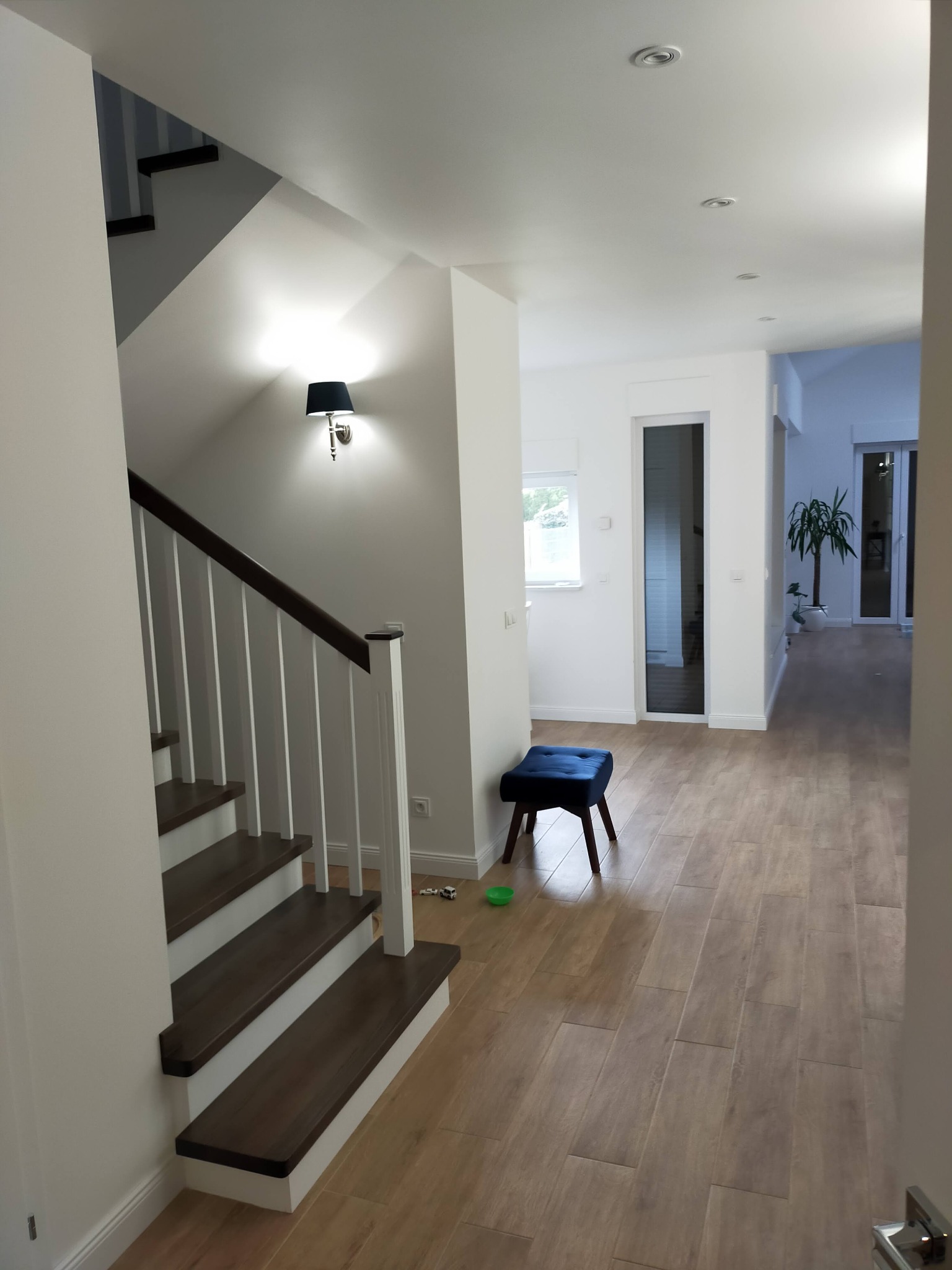 Nowoczesne wnętrze domu z drewnianymi schodami z białymi balustradami, podłogą z paneli imitujących drewno, niebieskim pufem i dekoracyjną lampą ścienną.