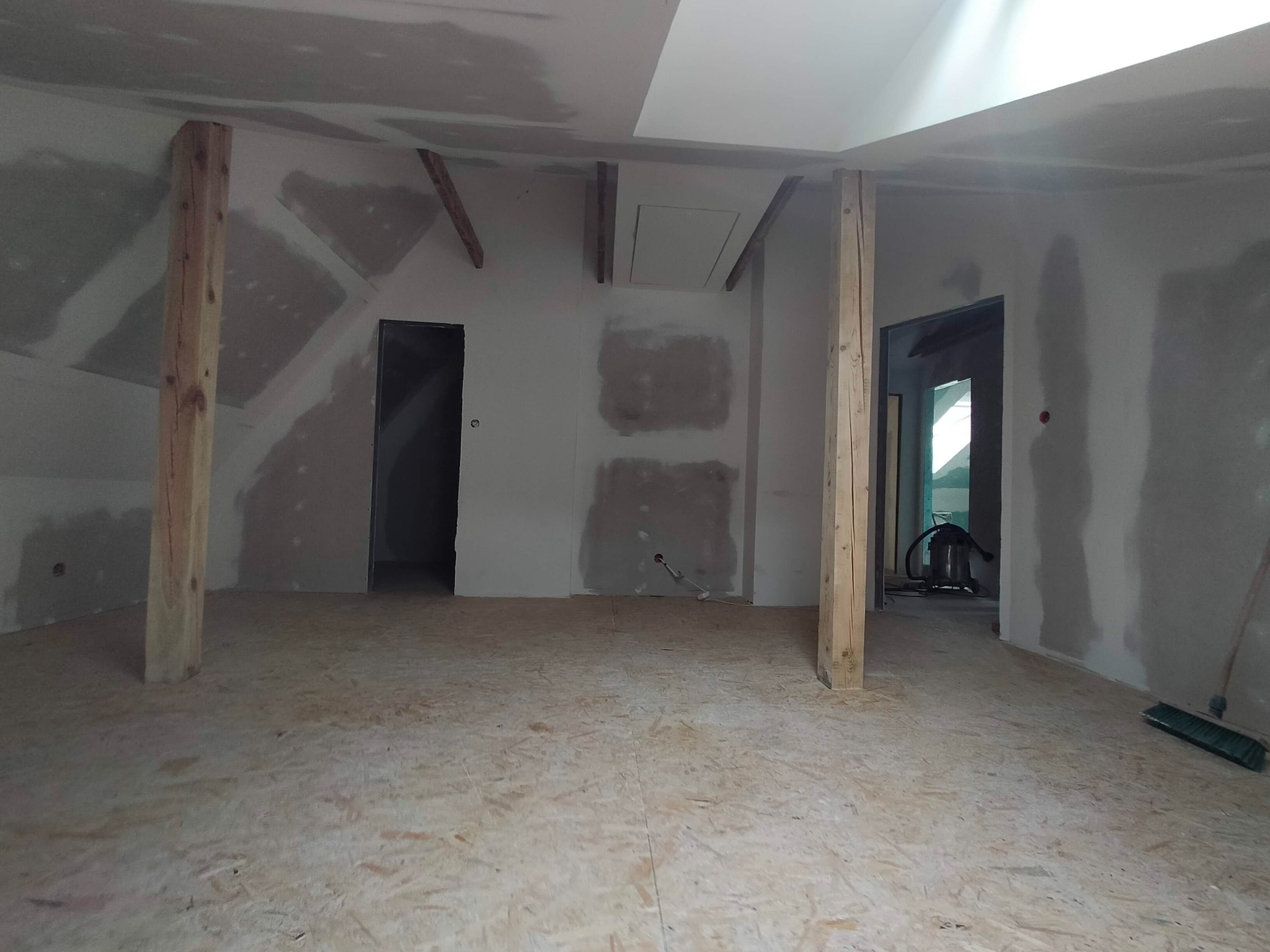 Poddasze w trakcie prac wykończeniowych: surowe drewniane słupy, płyty kartonowo-gipsowe z widocznymi miejscami szpachlowania, podłoga z płyt OSB i otwarte przejście do sąsiedniego pomieszczenia...