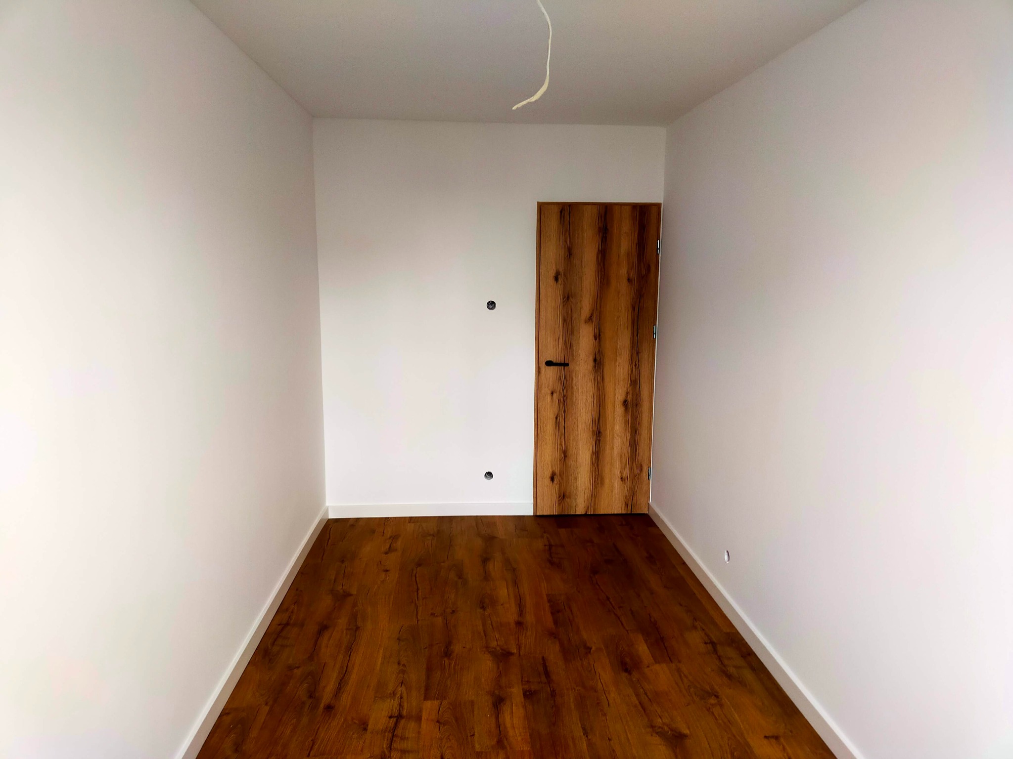 Pusty pokój po remoncie z panelami podłogowymi w odcieniach brązu i drewnianymi drzwiami z czarną klamką, białe ściany i listwy przypodłogowe, widoczny kabel wystający z sufitu.