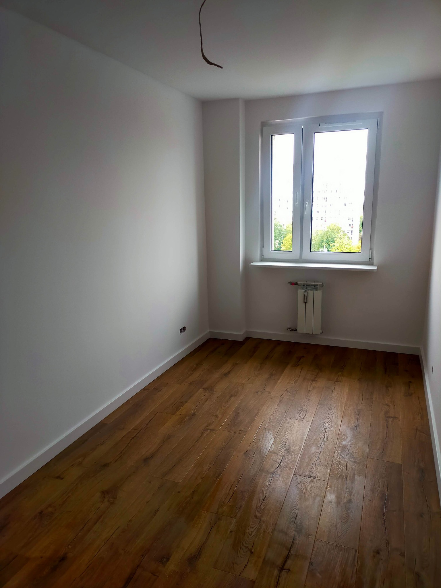 Pusty pokój po remoncie: świeżo pomalowane białe ściany, brązowe panele podłogowe, białe okno z widokiem na zieleń. Widoczny grzejnik pod oknem i wystający kabel ze ściany.