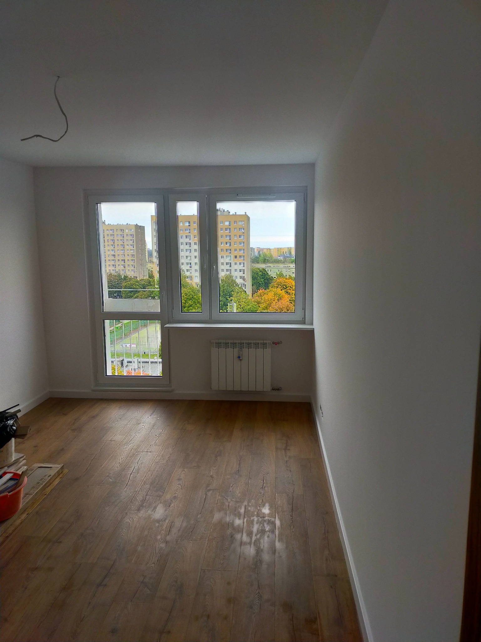 Pusty pokój po remoncie z drewnianą podłogą, świeżo pomalowanymi ścianami i widokiem na blok zza okna.