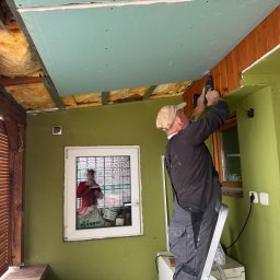 Usługi budowlane DARO - Monter na drabinie przykręca płytę gipsowo-kartonową do drewnianej konstrukcji sufitu w pomieszczeniu z zielonymi ścianami i oknem z luksferami.