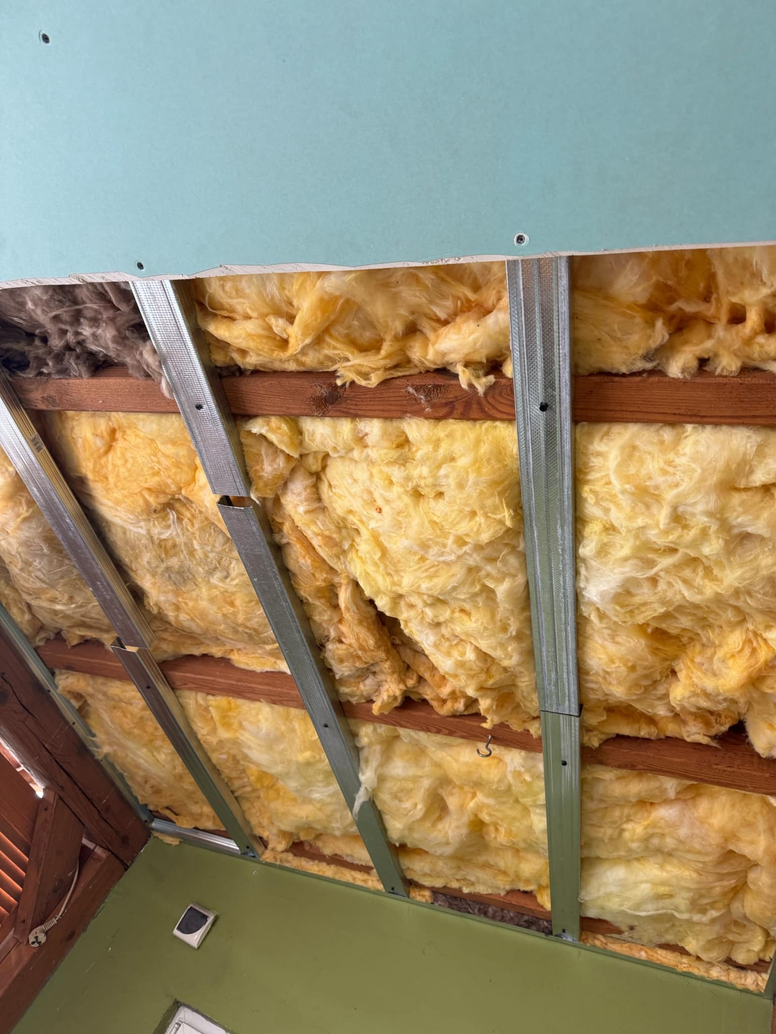 Ocieplenie poddasza wełną mineralną, widoczne metalowe profile, drewniana konstrukcja i fragment płyty gipsowej.