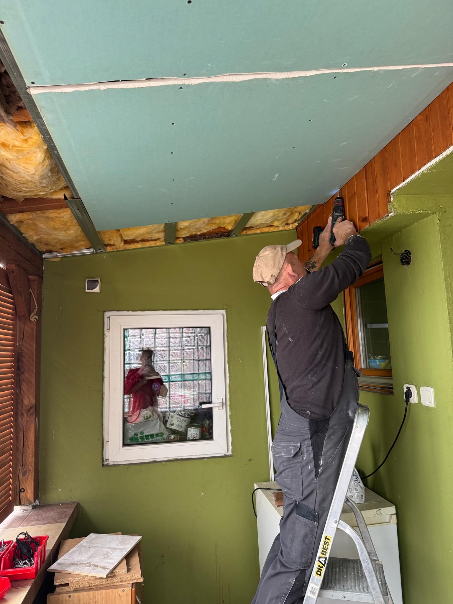 Monter na drabinie przykręca płytę gipsowo-kartonową do drewnianej konstrukcji sufitu w pomieszczeniu z zielonymi ścianami i oknem z luksferami.