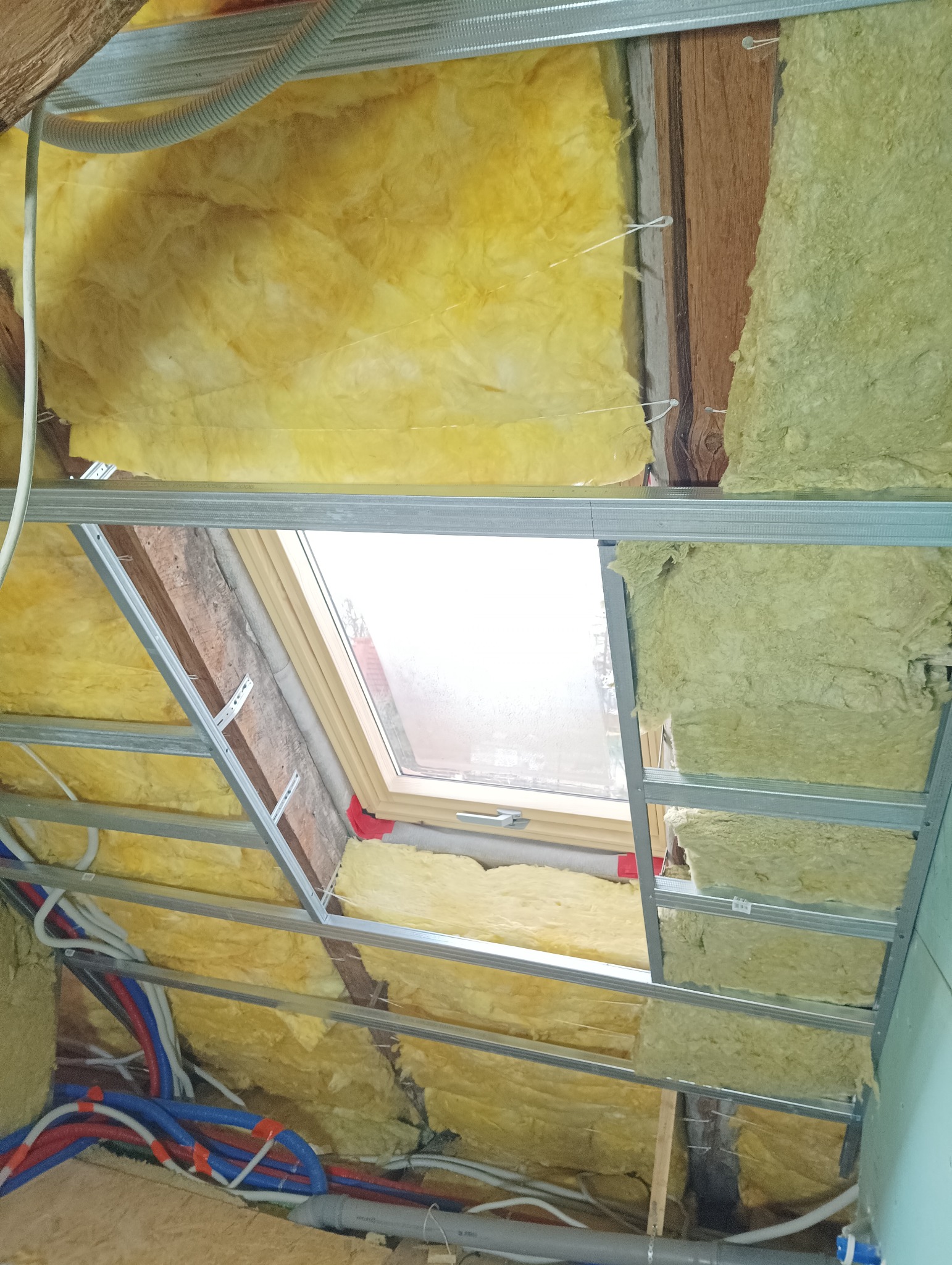 Izolacja poddasza wełną mineralną z widoczną konstrukcją metalową i oknem dachowym, instalacje wodne i kanalizacyjne w tle.