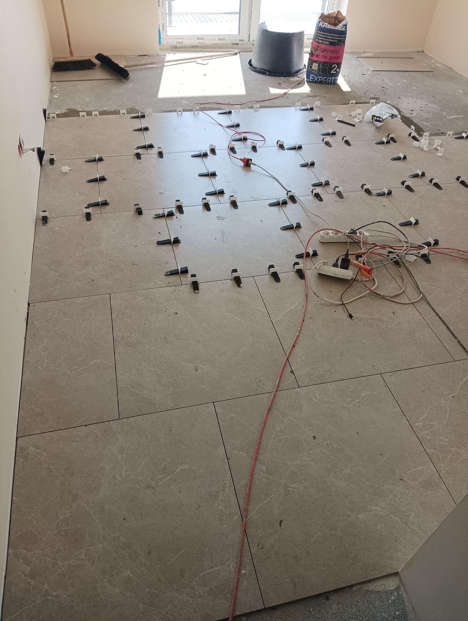 Układanie płytek podłogowych w trakcie realizacji, widoczne kliny poziomujące, szpachla, wiadro i worek kleju w tle.