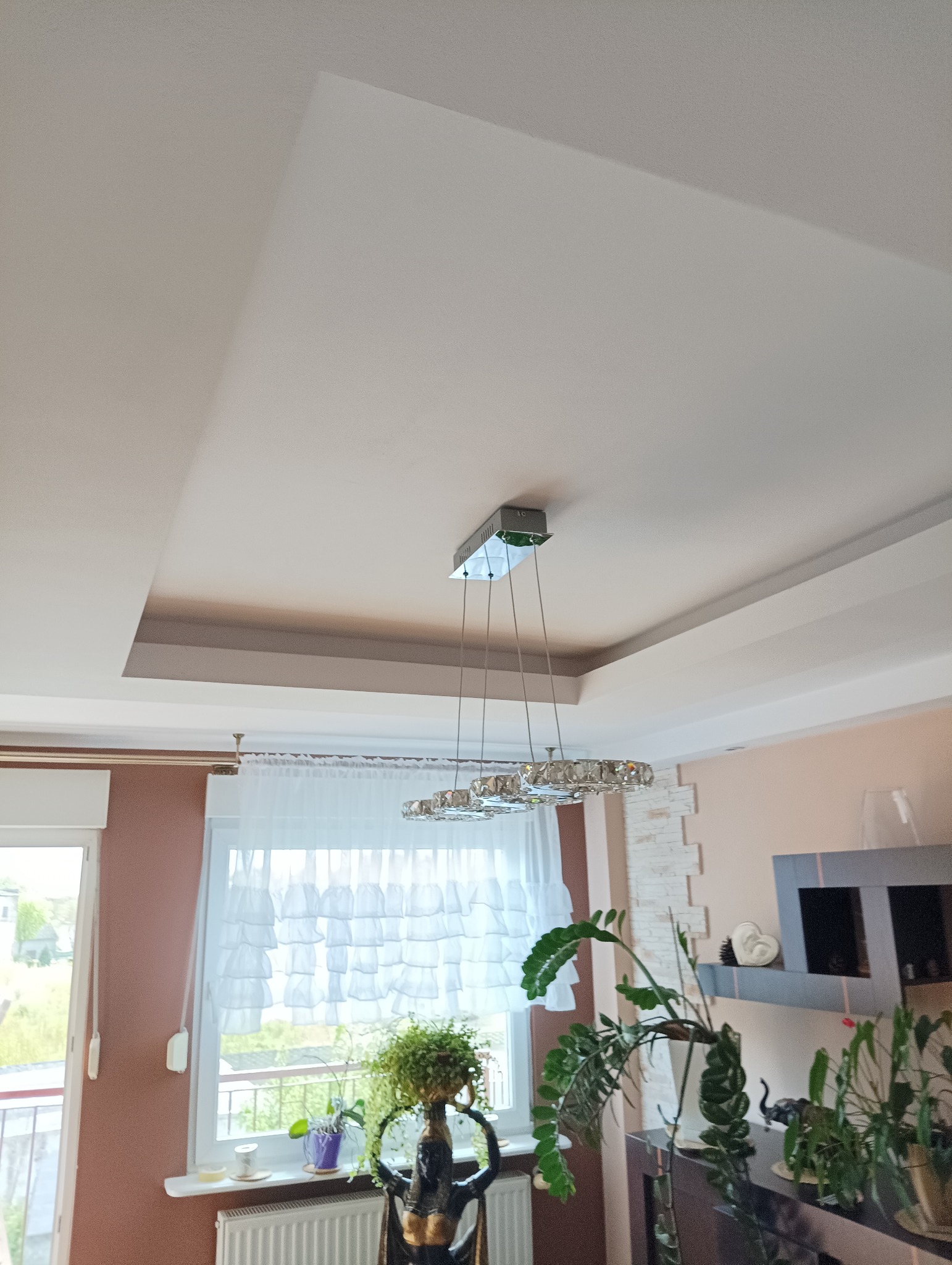 Wykończony sufit podwieszany z dekoracyjnym oświetleniem kryształowym w salonie z widokiem na okno i rośliny doniczkowe.