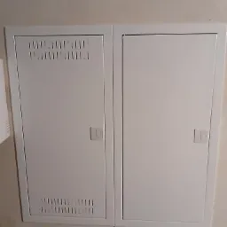 Biała, podwójna skrzynka elektryczna z wentylacją na jednej z klap, zamontowana na ścianie wewnątrz budynku.