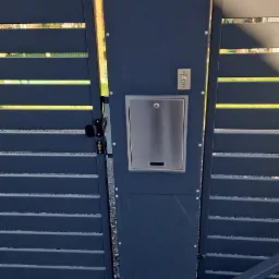 Nowoczesna, grafitowa brama wjazdowa z poziomymi panelami, wyposażona w skrzynkę na listy ze stali nierdzewnej i przycisk dzwonka, osadzona na żwirowym podłożu.