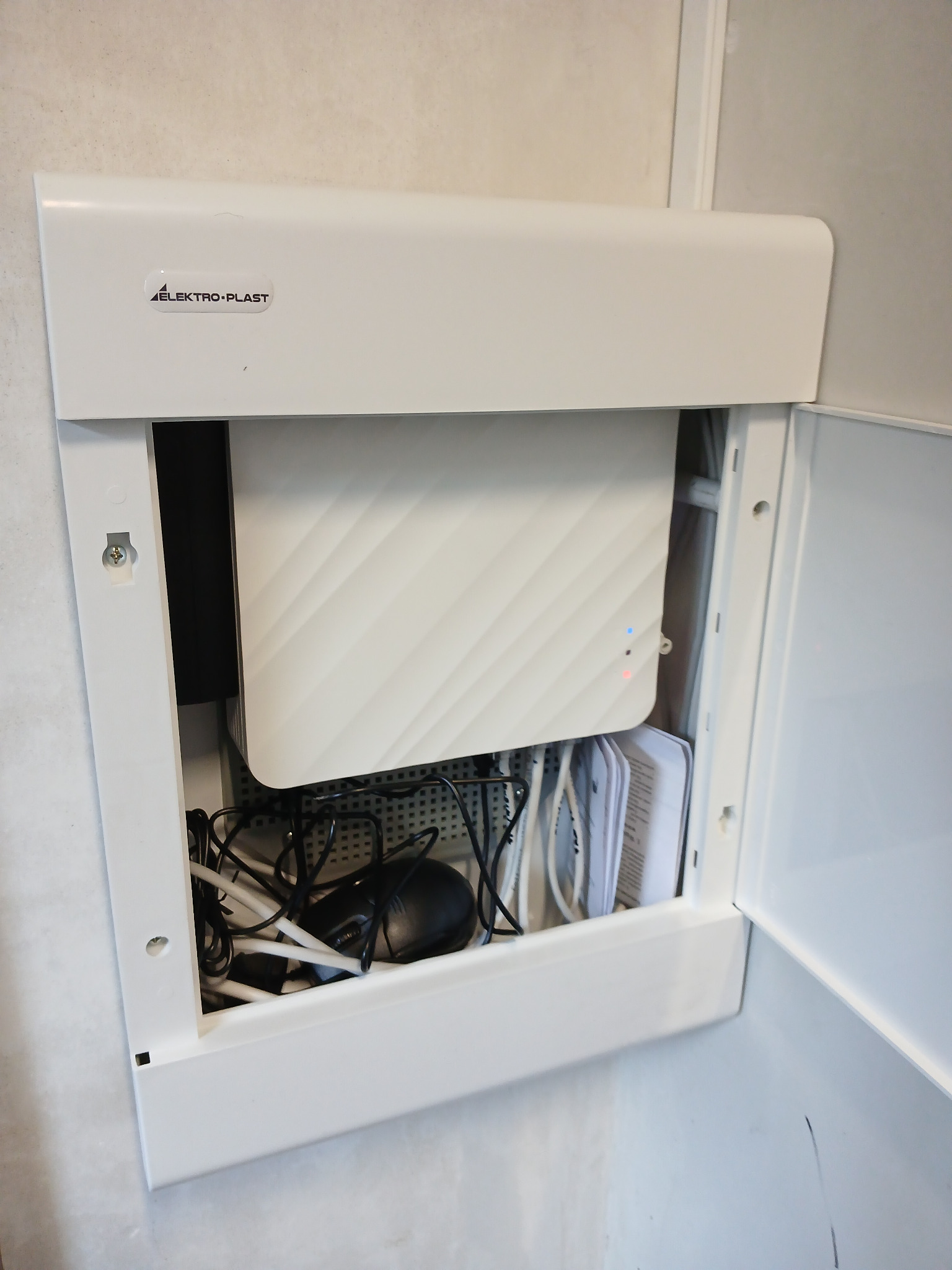 Otwarta, biała szafka elektryczna z widocznym routerem WiFi z diodami LED, przewodami i elementami montażowymi, marki Elektro-Plast.