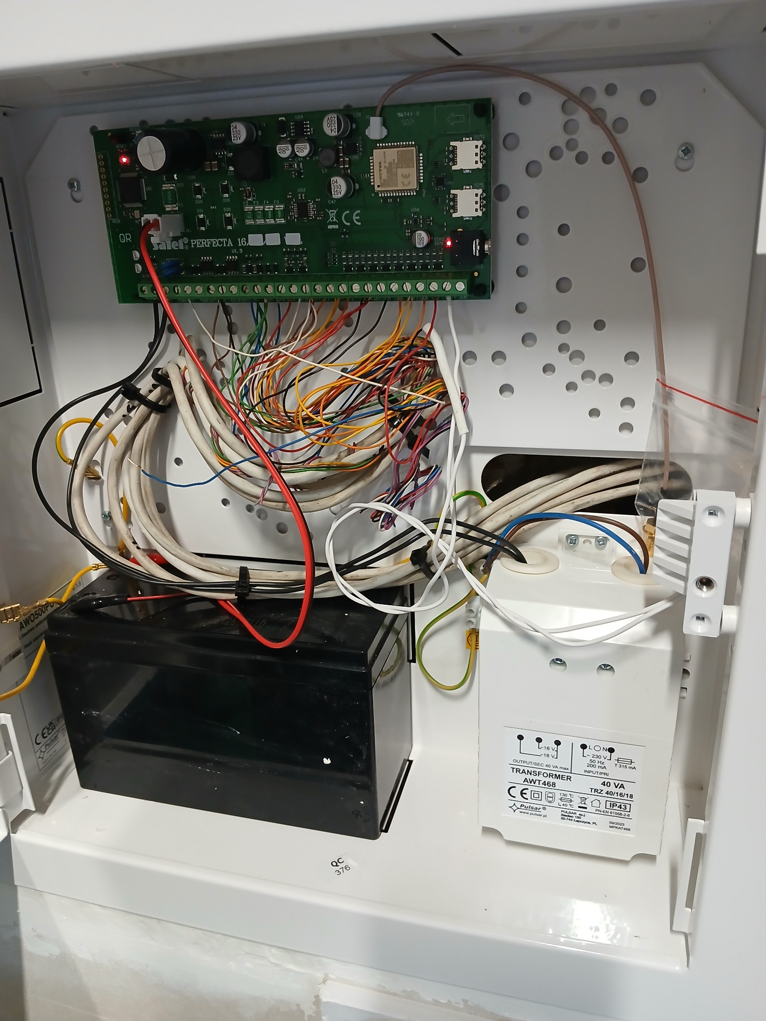 Wnętrze skrzynki z elektroniką systemu alarmowego: płyta główna Satel Perfecta 16, transformator Pulsar AWT468, akumulator, okablowanie. Widoczne złącza, diody LED i oznaczenia na podzespołach.