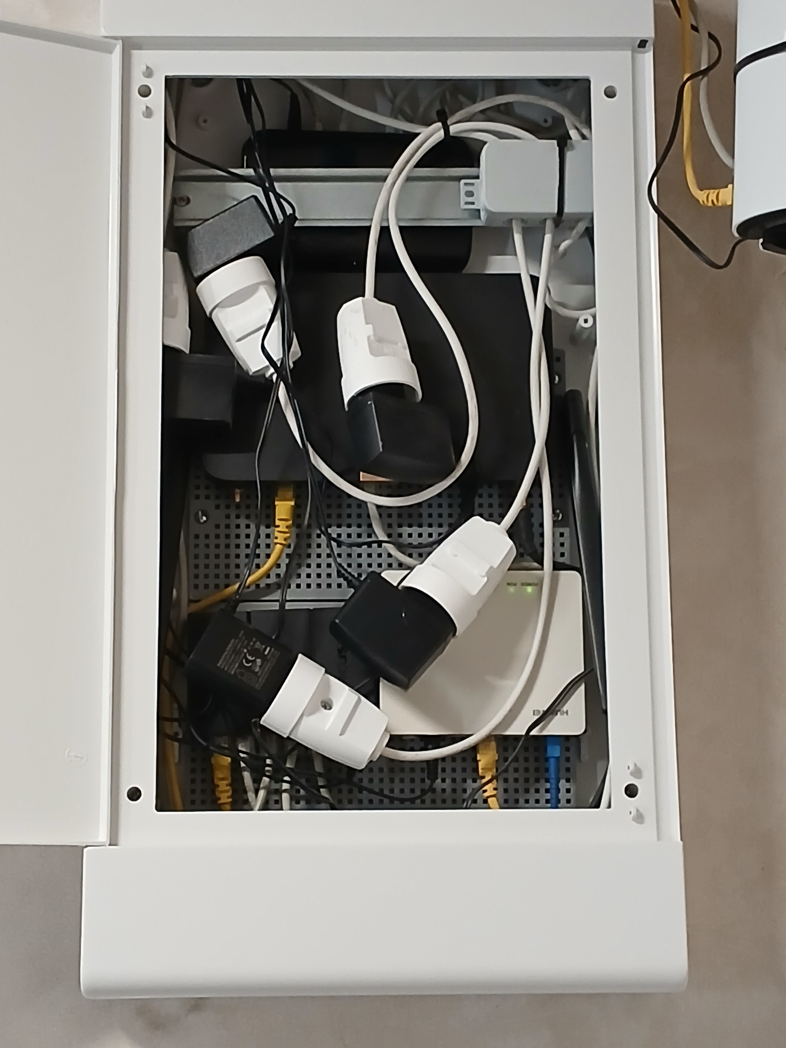 Otwarta biała szafka z widocznymi kablami, zasilaczami i routerem, zamontowana na ścianie, z żółtymi i niebieskimi kablami Ethernet.